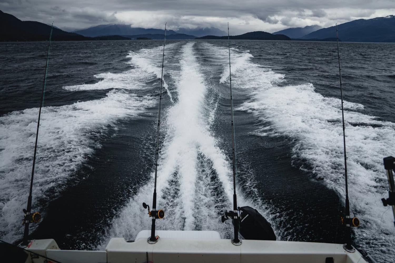 Trolling rods off the stern of a boat under way, wake trailing toward forested north-coast mountains